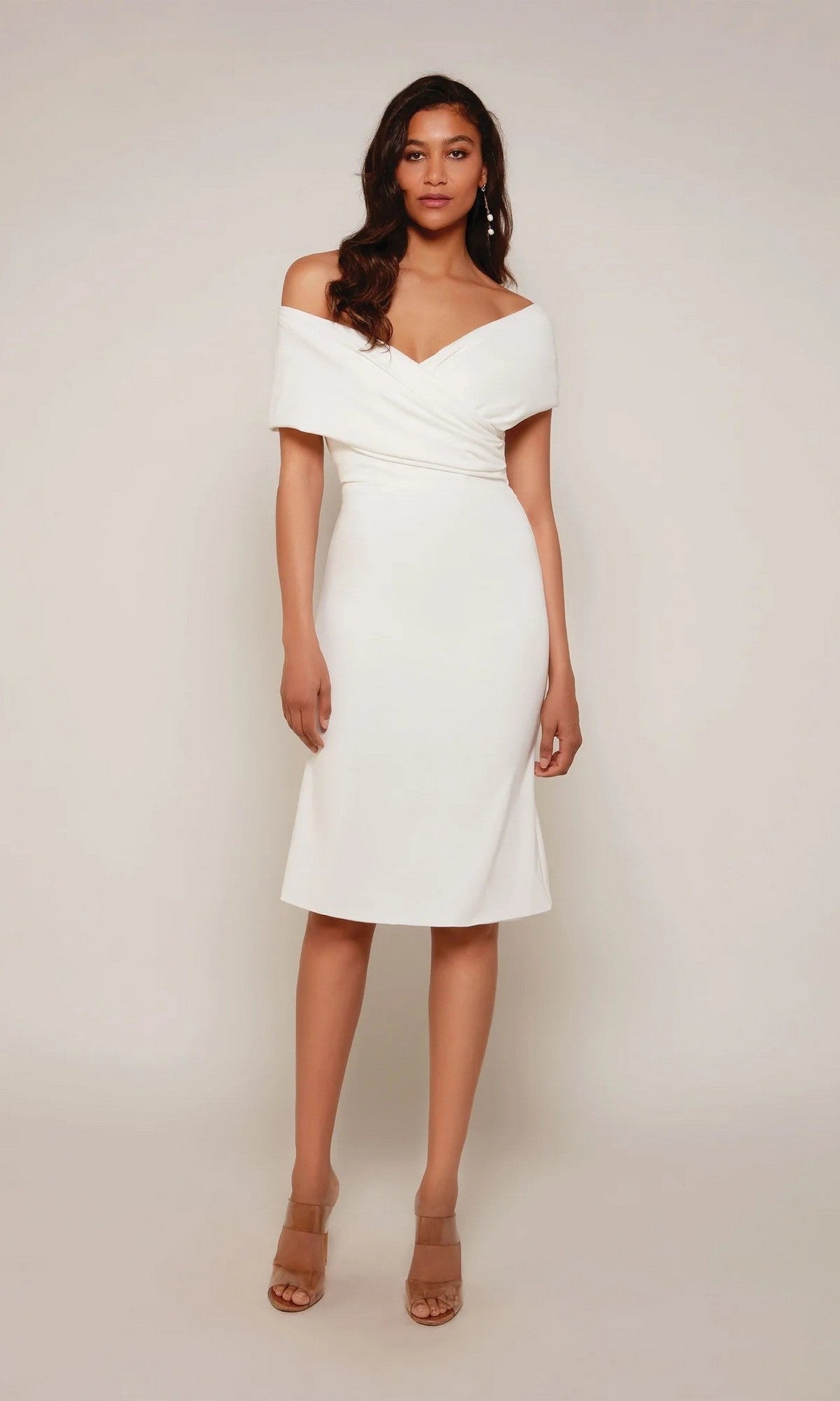 A woman faces forward in the Alyce 70023 knee-length white party dress with a classic sheath silhouette and beige heeled sandals, standing against a plain light background.